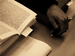 Das Bild zeigt eine Detailaufnahme: Neben einem aufgeschlagenen Buch ist die Hand des Lesers mit einem Kugelschreiber zu sehen.