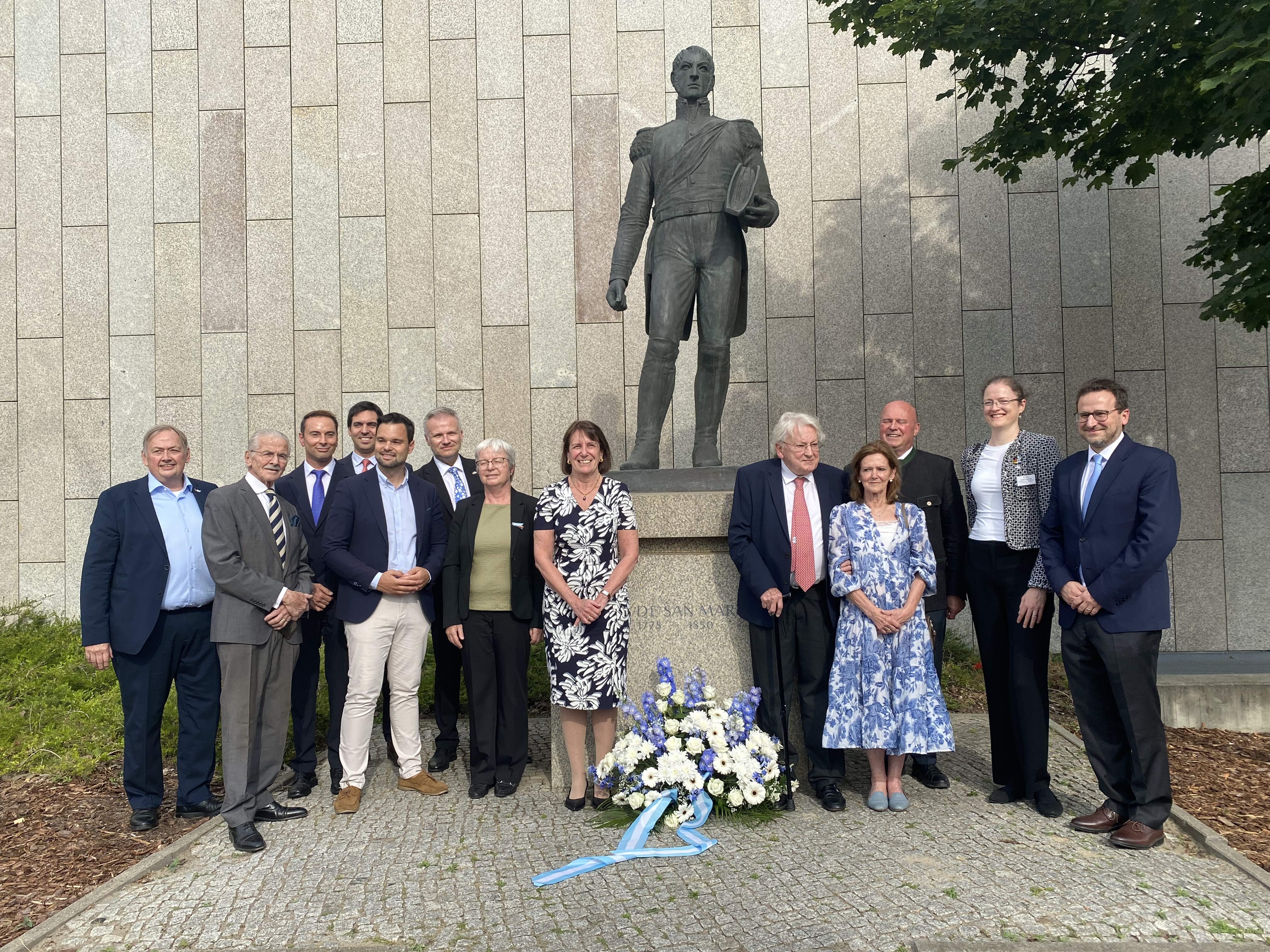 Personen und ein Blumenkranz vor einer Bronzestatue