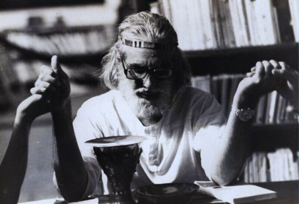 Ernesto Cardenal sitzt an einem Tisch und hält andere Personen (nicht im Bild) an den Händen