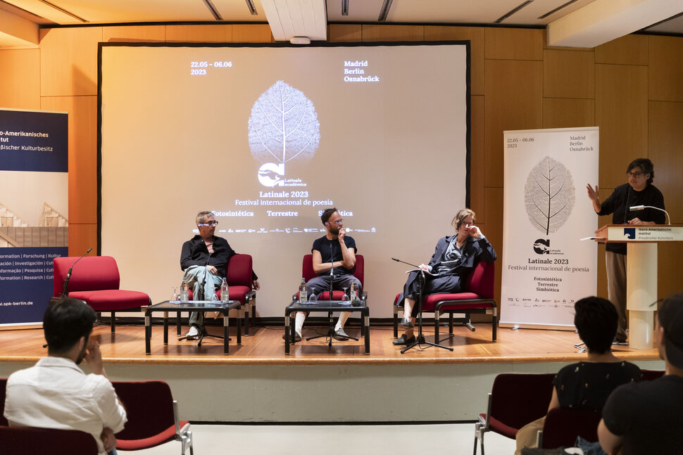 3 artistas no palco ouvem o apresentador, que fala em uma mesa de pé.