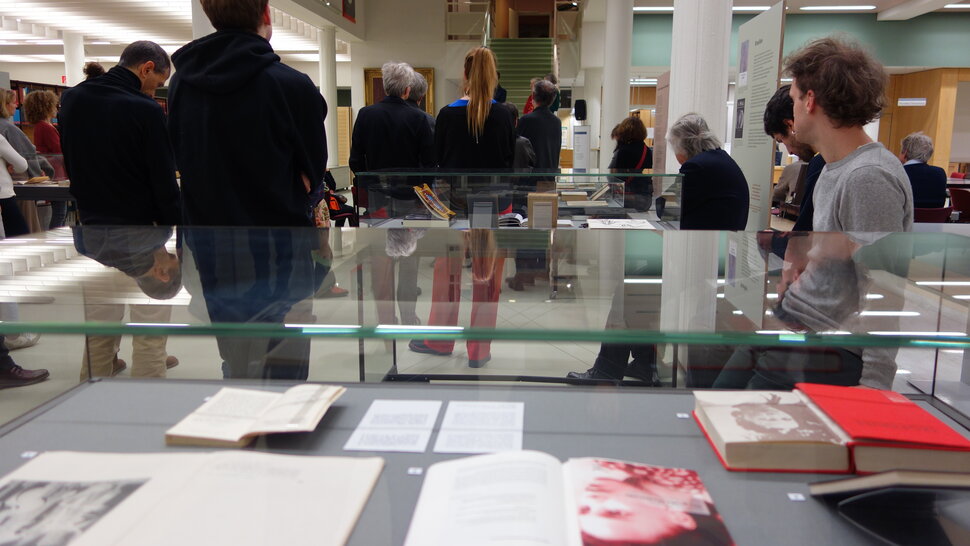 As pessoas ouvem a introdução durante a abertura da exposição, em frente a uma vitrine com livros (abre uma ampliação da imagem)