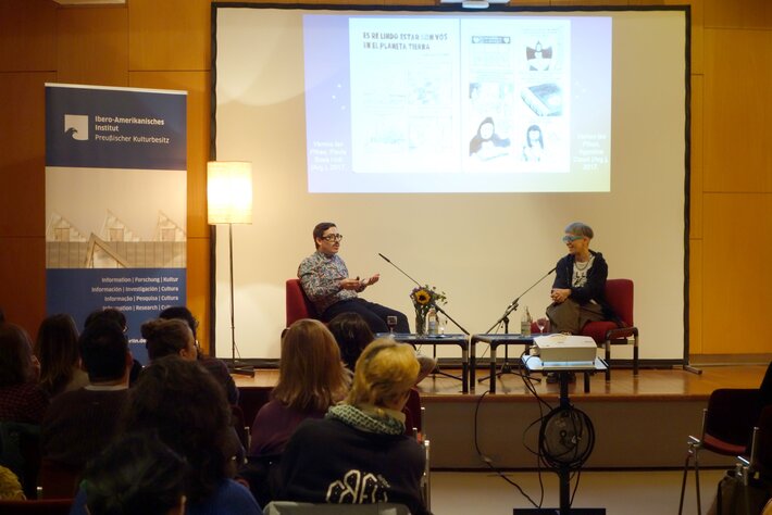 Powerpaola und Amadeo Gandolfo im Gespräch auf der Bühne; im Hintergrund Projektion aus einem Comic (öffnet Vergrößerung des Bildes)