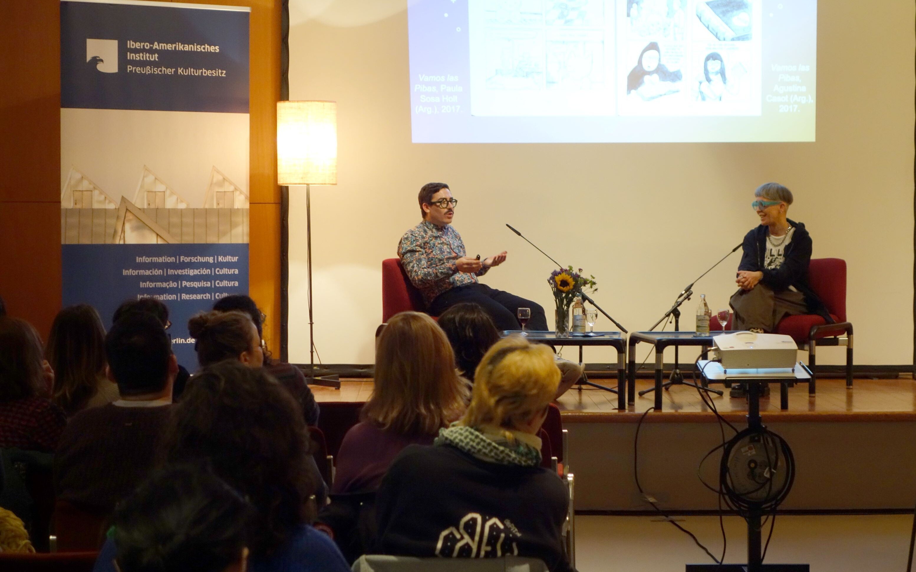 Powerpaola e Amadeo Gandolfo em conversa no palco; projeção de uma história em quadrinhos ao fundo