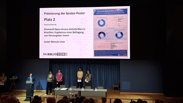 Personas de pie en un escenario frente a una gran diapositiva sobre premiación de pósteres, con público sentado.