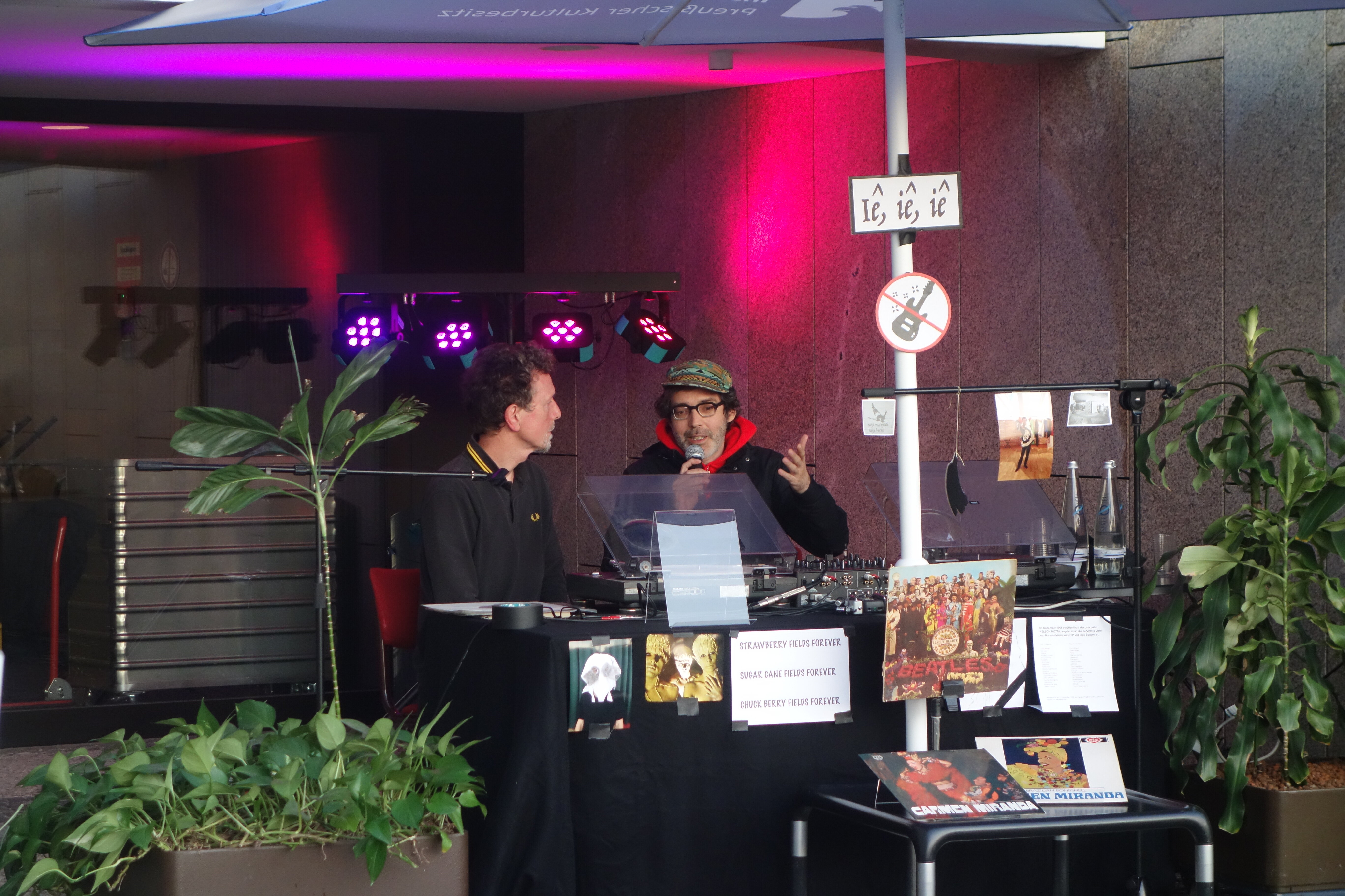 DJ detrás de la mesa de mezclas, con discos, fotos y palmeras al borde (abre una ampliación de la imagen)