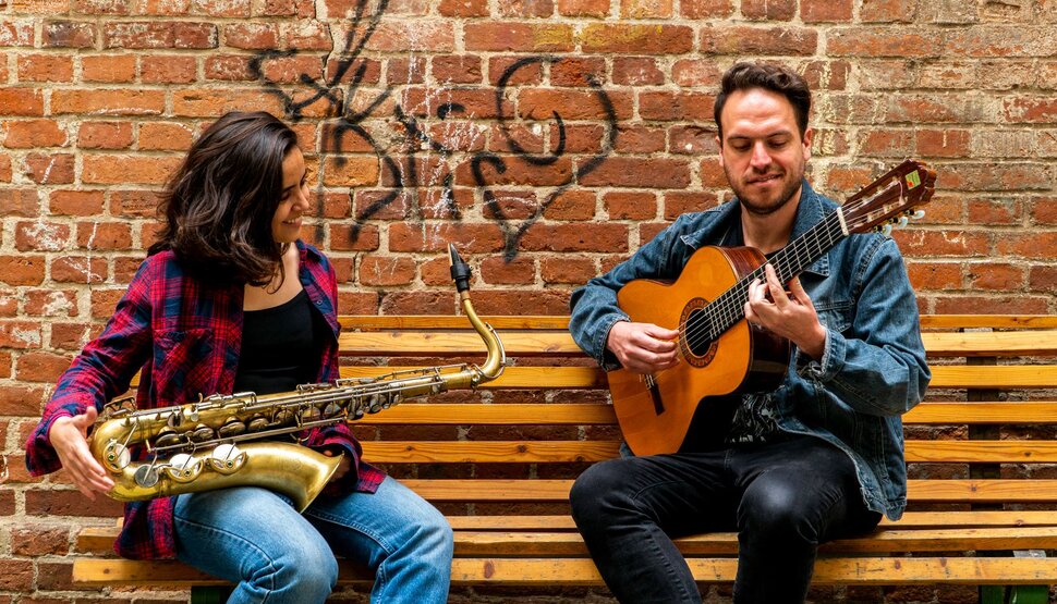 Die beiden Musiker sitzen nebeneinander auf einer Bank und musizieren (öffnet Vergrößerung des Bildes)