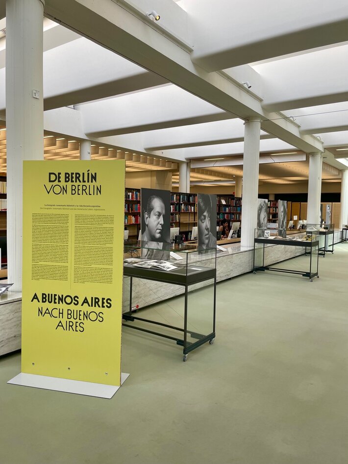 Blick in die Ausstellung im Lesesaal mit Fotoporträts und Vitrinen, vorn eine hohe Tafel mit den Ausstellungsdaten (öffnet Vergrößerung des Bildes)