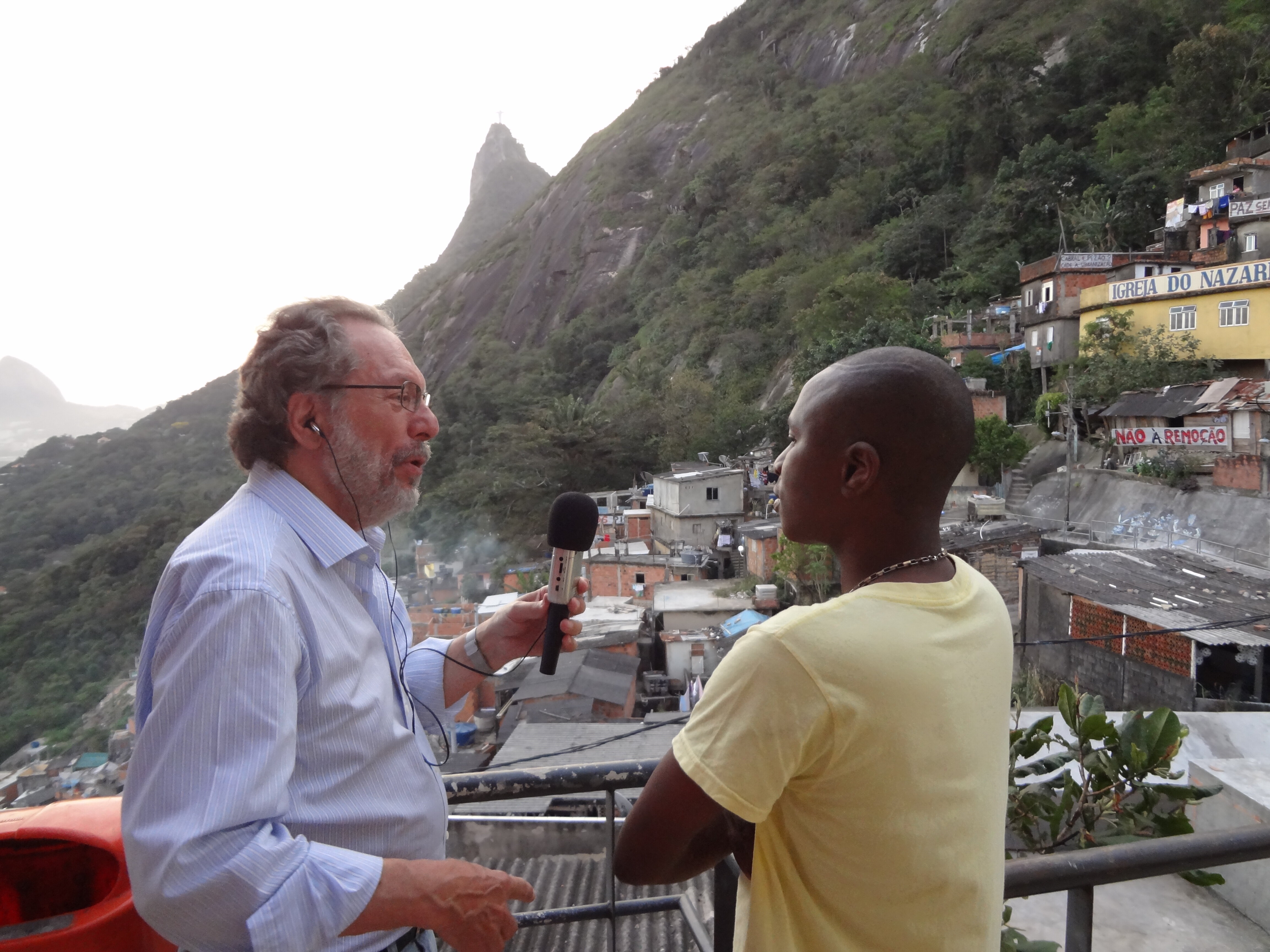 Ein Mann trägt Kopfhörer und Mikrofon und interviewt einen anderen Mann vor dem Hintergrund einer Favela in Rio de Janeiro (öffnet Vergrößerung des Bildes)