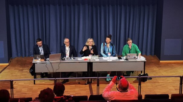 Mesa redonda con cinco personas sentadas, una persona hablando, público en primer plano.