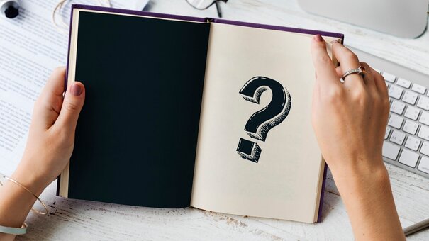 Hände halten ein aufgeschlagenes Notizbuch, auf dessen einer Seite ein Fragezeichen steht.