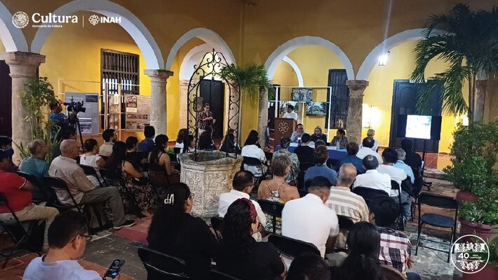 Ein sitzendes Publikum hört einem Vortrag zu, Ort ist ein Innenhof eines gelben Gebäudes mit Kolonnadengang