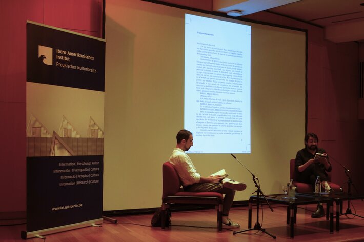 Autor und Moderator auf der Bühne; der Autor liest vor, im Hintergrund wird sein Roman projiziert. (öffnet Vergrößerung des Bildes)
